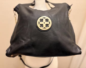 Könnte beinhalten: Eine schwarze Handtasche aus Kunstleder mit strukturierter Oberfläche und einem markanten, kreisförmigen Emblem aus goldfarbenem Metall in der Mitte der Vorderseite. Die Tasche hat schwarze Trageriemen und goldfarbene Beschläge.