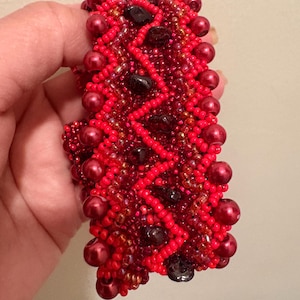 May include: A wide bracelet crafted with vibrant red seed beads arranged in a zigzag pattern, accented with larger, glossy red spherical beads and interspersed with dark, irregular-shaped beads throughout the intricate, textured beadwork design.