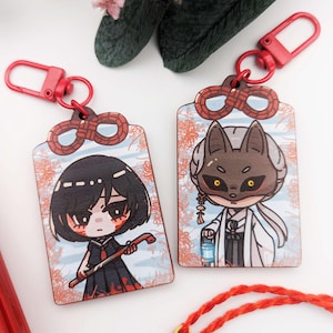 May include: Two rectangular keyrings with red details. One shows a cartoon girl with black hair and a staff, the other a fox-like character. Both have red clasps and floral patterns in the background.