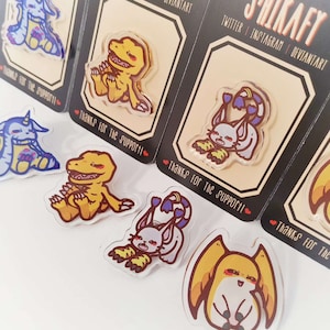 May include: Acrylic pins featuring cartoon characters in various poses. The pins are on black backing cards with the text "THANKS FOR THE SUPPORT!" and the artist's social media handles. The characters are in shades of yellow, blue, and white.