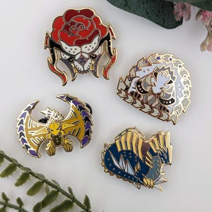 Può includere: Quattro spille smaltate con finiture color oro. Le spille presentano vari disegni, tra cui una rosa rossa, una creatura bianca e grigia, una creatura alata gialla e viola e una creatura blu e oro.