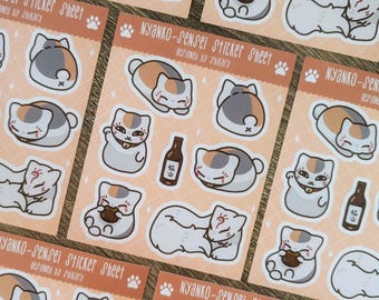 以下が含まれることがあります： 額に赤い印のある三毛猫のイラストが描かれたステッカーシート9枚セット。猫は座ったり寝たり、酒の瓶を持ったりと様々なポーズをとっています。ステッカーシートは白地に桃色の背景で印刷されており、「NYANKO-Sensei Sticker Sheet」の文字が印刷されています。