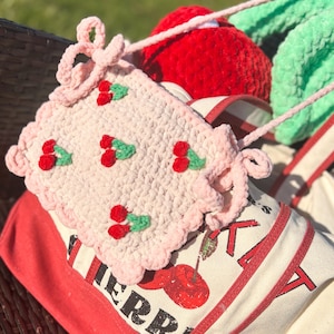 May include: A pink crocheted bag with a cherry design. The bag has a long strap and is sitting on a red and white striped tote bag.