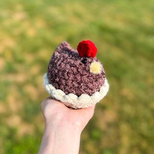 May include: A handmade, plush cupcake toy with a dark purple body, a red cherry on top, and a cream-colored base. The cupcake has a yellow dot and a black bead for the eye. The toy is held in a hand against a blurred green background.