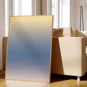 Puede incluir: Una impresión de arte abstracto enmarcada con un degradado de colores azules y amarillos. La impresión se apoya en un sofá beige claro.