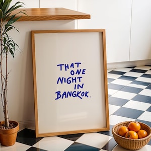May include: A framed print with the text "THAT ONE NIGHT IN BANGKOK" in blue handwritten style. The print is in a light wood frame, leaning against a wall. A potted plant and a basket of oranges are in the foreground.