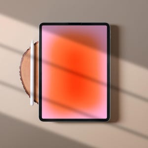 Puede incluir: Una tableta digital con una pantalla degradada naranja y rosa vibrante, acompañada de un lápiz óptico blanco. La tableta descansa sobre una rodaja de madera, con sombras proyectadas sobre una superficie de tono neutro.