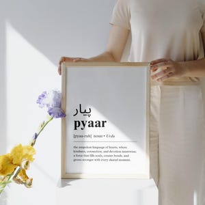 May include: A framed print with the word "pyaar" in black text, defined as the unspoken language of hearts in Urdu. The print is on a white background and is surrounded by a light wood frame.