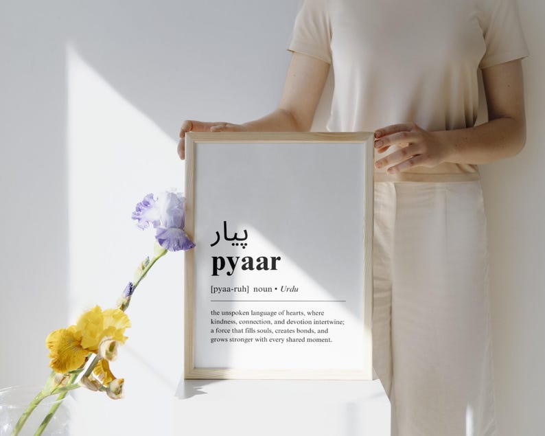 May include: A framed print with the word "pyaar" in black text, defined as the unspoken language of hearts in Urdu. The print is on a white background and is surrounded by a light wood frame.