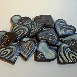 Pode incluir: Uma coleção de ornamentos de cerâmica em forma de coração com vários desenhos, incluindo espirais, padrões de dente-de-leão e a palavra "LOVE". Os corações são pintados em tons de azul e branco sobre um fundo escuro.