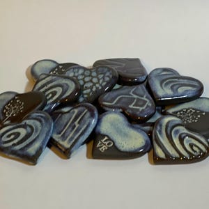 Pode incluir: Uma coleção de pequenos azulejos de cerâmica em forma de coração com vários padrões azuis e brancos. Alguns dos corações têm um design de espiral, enquanto outros têm um padrão mais abstrato. Um coração tem a palavra "LOVE" escrita nele.