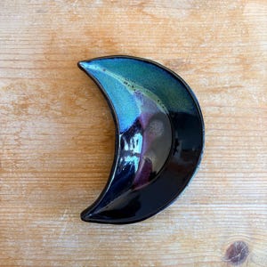 May include: A ceramic crescent-shaped dish with a glossy finish. The dish has a gradient of teal, blue, and purple, with a black base. The dish is on a wooden surface. This decorative dish is ideal for holding small items.