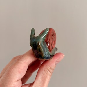May include: A small ceramic snail figurine featuring a glossy, multi-toned body with blended shades of blue, green, and black, paired with a distinct, rounded shell finished in a vibrant, deep red glaze with a smooth, reflective surface texture.