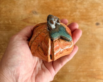 May include: A small, hand-sculpted ceramic pumpkin figurine with a glossy orange glaze and a textured surface. The pumpkin features a prominent, dark brown and teal-coloured stem detail on top, showcasing a smooth, reflective finish throughout.