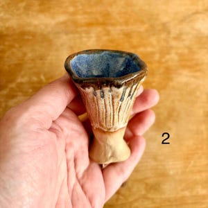 May include: A small ceramic cup with a flared, uneven rim and a tapered base. The interior features a deep blue glaze, while the exterior displays a textured, light tan finish with vertical brown drip patterns extending from the rim downwards.