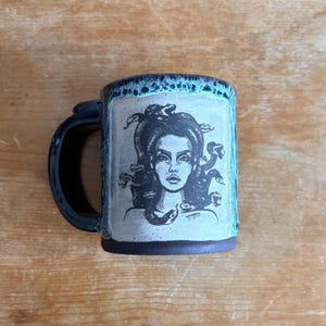 May include: A ceramic mug with a dark handle and base, featuring a light-coloured central panel with a black ink illustration of a figure with snakes for hair. The rim and handle area have a textured, mottled teal and dark blue glaze finish.