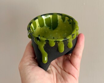 Puede incluir: Una pequeña taza de cerámica con exterior negro mate e interior verde lima brillante. El esmalte verde gotea sobre el borde superior, creando un efecto texturizado y superpuesto que recorre los lados del recipiente oscuro y redondeado.