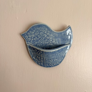 May include: A bird-shaped ceramic wall pocket featuring a glossy light blue glaze with a white floral and botanical pattern. The piece has a curved opening at the centre, creating a functional pocket for small items or decorative displays.