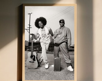 May include: Black and white photograph of two men standing in front of a brick wall. One man is wearing a white shirt and a bow tie, and the other man is wearing a black jacket and a baseball cap. A guitar is leaning against the wall.