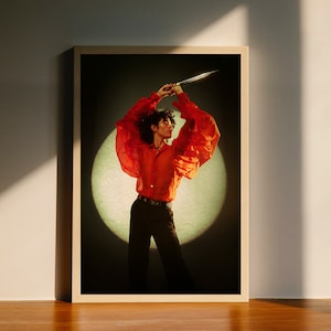 Könnte beinhalten: Eine Person in einem roten, durchsichtigen, gekräuselten Hemd hält ein Messer vor einem Scheinwerfer. Sie trägt eine schwarze Hose und einen schwarzen Gürtel.