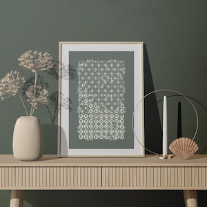Puede incluir: Una impresión enmarcada con un patrón geométrico en blanco sobre un fondo verde salvia. La impresión está sobre una mesa de madera con un jarrón de flores secas, un candelero y una concha.