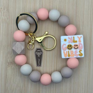 May include: A beaded bracelet keychain with a circular design. The bracelet features a mix of pink, grey, and white beads, with a wooden cube accent. It includes a gold-coloured clasp and a charm with the words "ONLY GOOD VIBES". A coffin charm is also included.