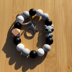 May include: A beaded keychain bracelet featuring a raccoon charm. The bracelet is made of black, white, and speckled gray beads. A silver-colored clasp and key ring are attached. A small wooden geometric shape is also included.