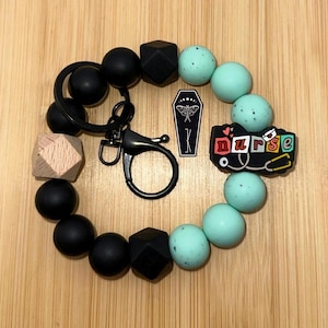 May include: A black beaded keychain wristlet with a lobster clasp. The wristlet features black, mint green, and wooden beads, along with a charm that says "nurse" and a coffin charm with a moth design.