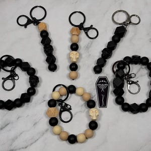 May include: A collection of three beaded wristlet keychains and two vertical beaded keychains featuring black, tan, and wood-toned beads, including skull-shaped beads and black metal hardware. A small black coffin-shaped charm with a white moth graphic is included.