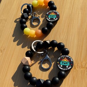 May include: Two black beaded wrist keyrings with a rainbow gradient of yellow, orange, and black beads. Each keyring has a round charm with the text "THIS LITTLE LIGHT OF MINE I'M GONNA LET IT SHINE".