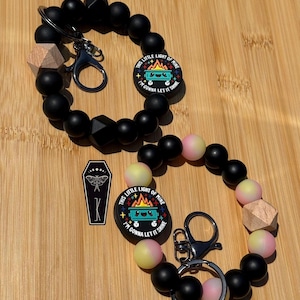 May include: Two circular wristlet keychains featuring matte black silicone beads, one wooden geometric bead, and a round charm displaying a cartoon dumpster fire with the text This Little Light Of Mine I'm Gonna Let It Shine. One includes a coffin charm.
