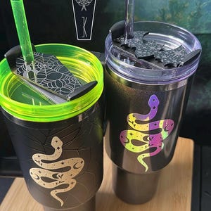 May include: Two black tumblers with lids and straws. The left tumbler has a neon green rim, a coffin-shaped topper with a spiderweb design, and a gold snake graphic. The right tumbler has a purple straw, a bat-shaped topper, and a holographic snake.