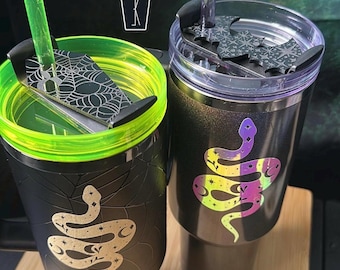 May include: Two black tumblers with lids and straws. The left tumbler has a neon green rim, a coffin-shaped topper with a spiderweb design, and a gold snake graphic. The right tumbler has a purple straw, a bat-shaped topper, and a holographic snake.