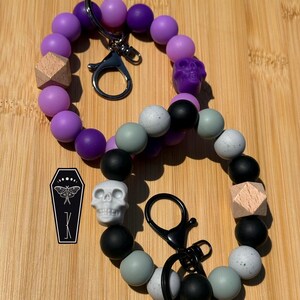 May include: Two circular silicone bead keychains with metal clasps. One features purple beads, a wooden geometric bead, and a purple skull bead. The other has black, grey, and speckled beads, a wooden geometric bead, and a white skull bead.
