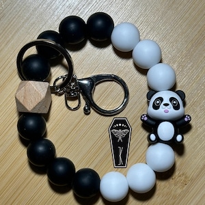 May include: Black and white silicone beaded keychain with a wooden bead and a panda charm. The keychain has a silver metal ring and clasp.