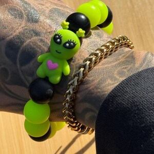 May include: A beaded bracelet featuring alternating neon green and black spherical beads with a central green alien charm that has a small pink heart on its chest, paired with a gold-toned metal chain bracelet with a woven, interlocking link design.