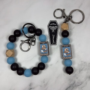 Peut inclure: Un ensemble de porte-clés en perles avec des perles en silicone noir mat et bleu clair, une perle en bois naturel et une breloque rectangulaire représentant un crâne sur une pile de livres avec le texte THE READER. Inclut une breloque cercueil.