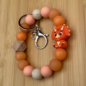 May include: A circular keychain bracelet featuring a mix of round silicone beads in shades of peach, orange, and speckled cream, along with a single hexagonal wooden bead. A silver metal keyring and lobster clasp are attached to the bracelet.