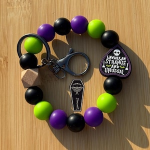 May include: A beaded keychain with black, purple, and neon green beads. The keychain includes a metal clasp, a wooden cube, and two charms. One charm is a black coffin with a moth, and the other is a purple heart with the text "I myself am strange and unusual."