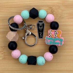 May include: A beaded keychain bracelet featuring round silicone beads in black, light pink, and mint green, along with a natural wood bead. It includes a silver metal clasp, a black coffin-shaped charm with a moth graphic, and a pink ER sign.