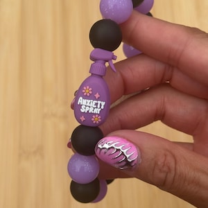 May include: A beaded bracelet featuring alternating matte black and glittery purple spherical beads. A central charm shaped like a purple spray bottle is included, featuring the text ANXIETY SPRAY in white letters, accented with small pink flower and star icons.