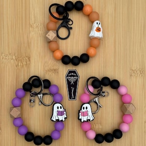 May include: Three Halloween wristlet keyrings. One is orange and black with a ghost holding a pumpkin. The other two are purple and pink with ghosts reading books. Each has a wooden bead and a metal clasp.