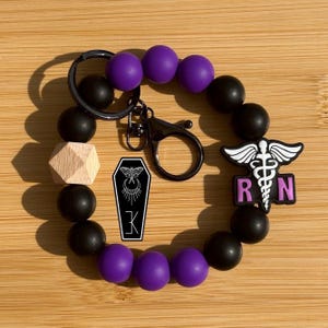 May include: A purple and black beaded bracelet with a silver keychain and a charm featuring a caduceus and the letters "RN". The charm is pink. There is also a wooden bead and a small black and white coffin charm with a moth design.