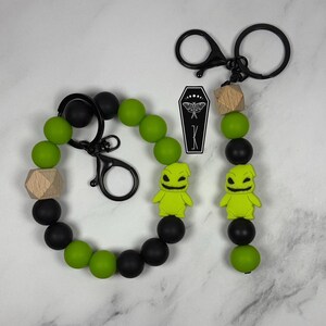 May include: A beaded keychain set featuring round matte black and lime green silicone beads, a natural wood geometric bead, and a lime green character charm with a wide grin. The set includes a black metal lobster clasp and a black metal keyring.