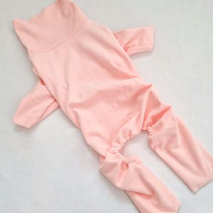 May include: A pale pink pet onesie with a high collar and long sleeves. The onesie has long legs with elasticated cuffs at the ankles. The garment is made of a soft, stretchy material.
