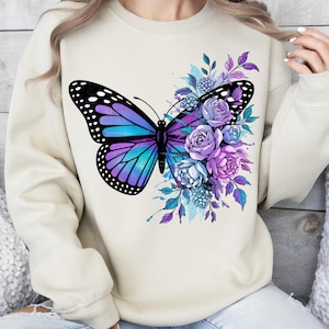 May include: A beige sweatshirt featuring a large butterfly design with purple and turquoise wings, and a cluster of purple and blue flowers. The butterfly has black polka dots on its wings. The design is centered on the front of the sweatshirt.