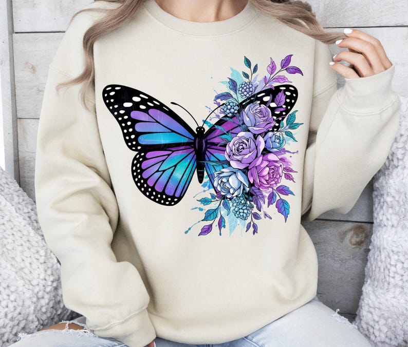 May include: A beige sweatshirt featuring a large butterfly design with purple and turquoise wings, and a cluster of purple and blue flowers. The butterfly has black polka dots on its wings. The design is centered on the front of the sweatshirt.