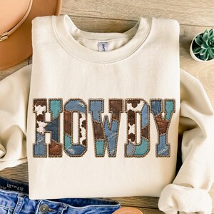 Puede incluir: Una sudadera de cuello redondo color crema con la palabra HOWDY en letras grandes. Las letras están rellenas con un patrón de retazos de estampado de vaca marrón y blanco, marrón sólido y tela azul claro con textura de mezclilla.