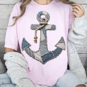 May include: A light pink short-sleeve t-shirt featuring a large, central graphic of a nautical anchor. The anchor is designed with a patchwork of blue denim and beige fabric textures, accented by a realistic rope illustration wrapped around the top.