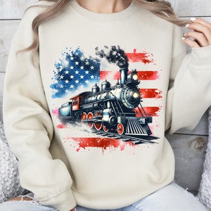 May include: A beige sweatshirt with a watercolor American flag design featuring a vintage steam locomotive. The locomotive is black and red with white smoke coming from the chimney.